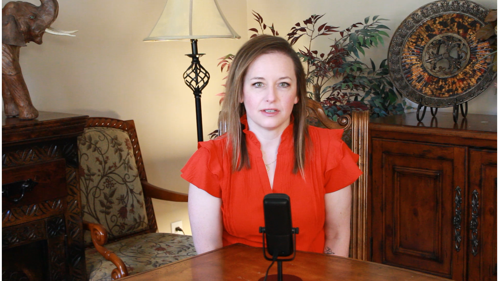 CEO Kellie in a red blouse sitting at a table with a microphone, in a room with wooden furniture and decorative items.